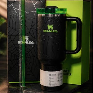 Peut inclure: Gobelet Stanley noir avec un couvercle et une poign&eacute;e verts, orn&eacute; d'un motif de toile d'araign&eacute;e. Le logo Stanley est en vert. Le gobelet est emball&eacute; avec une bo&icirc;te noire avec un motif de toile d'araign&eacute;e et une paille verte.