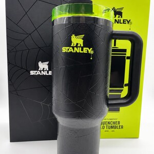 Peut inclure: Gobelet Stanley Quencher Flowstate noir de 1,18 L avec un motif toile d'araign&eacute;e. Le gobelet a une poign&eacute;e noire, un couvercle vert et une paille verte. Le logo Stanley est dor&eacute;. Il est &agrave; c&ocirc;t&eacute; d'une bo&icirc;te noire et d'une bo&icirc;te verte.