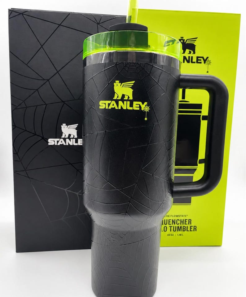 Peut inclure: Gobelet Stanley Quencher Flowstate noir de 1,18 L avec un motif toile d'araign&eacute;e. Le gobelet a une poign&eacute;e noire, un couvercle vert et une paille verte. Le logo Stanley est dor&eacute;. Il est &agrave; c&ocirc;t&eacute; d'une bo&icirc;te noire et d'une bo&icirc;te verte.