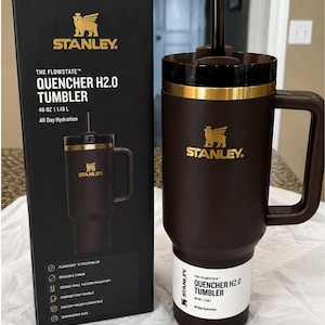 以下が含まれることがあります： ハンドルとストローが付いた茶色のスタンレーFlowstate Quencher H2.0タンブラー。黒い箱の隣にあります。タンブラーには金のバンドとスタンレーのロゴが付いています。箱には、再利用可能なストローや二重壁真空断熱材などの機能が記載されています。
