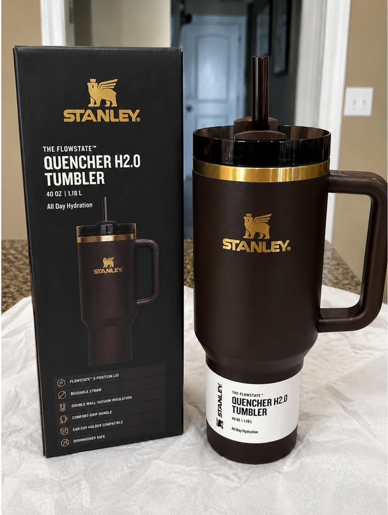 以下が含まれることがあります： ハンドルとストローが付いた茶色のスタンレーFlowstate Quencher H2.0タンブラー。黒い箱の隣にあります。タンブラーには金のバンドとスタンレーのロゴが付いています。箱には、再利用可能なストローや二重壁真空断熱材などの機能が記載されています。