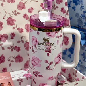 May include: White Stanley tumbler with a pink floral rose pattern, a pink lid, and a white handle. The tumbler has a gold band and a pink bow on the straw. The text "STANLEY" and "LoveShackFancy" are printed on the tumbler.