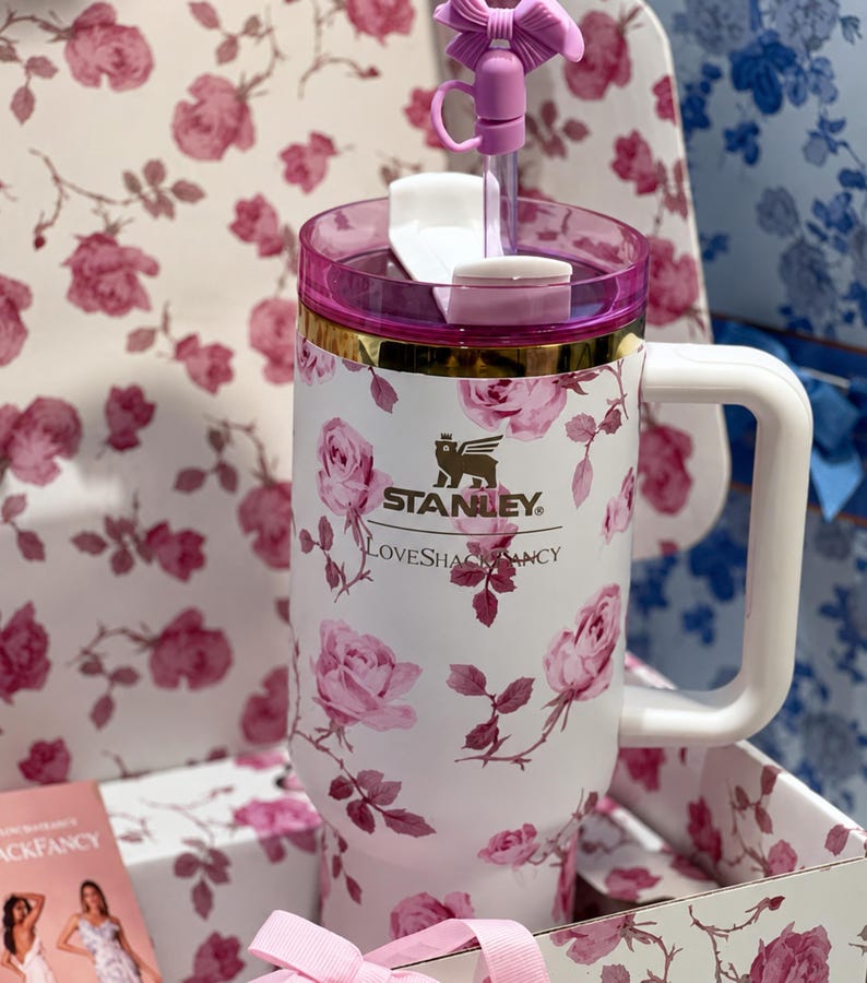 May include: White Stanley tumbler with a pink floral rose pattern, a pink lid, and a white handle. The tumbler has a gold band and a pink bow on the straw. The text "STANLEY" and "LoveShackFancy" are printed on the tumbler.