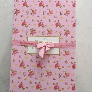 May include: A rectangular pink gift box with a floral pattern of pink and yellow roses. The box is wrapped with a pink ribbon that has the words "STANLEY" and "LOVESHACKFANCY" printed on it. A pink bow is tied on top.