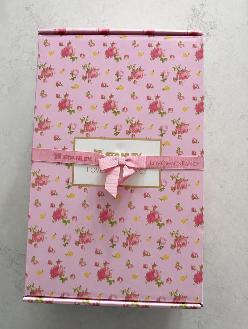 May include: A rectangular pink gift box with a floral pattern of pink and yellow roses. The box is wrapped with a pink ribbon that has the words "STANLEY" and "LOVESHACKFANCY" printed on it. A pink bow is tied on top.