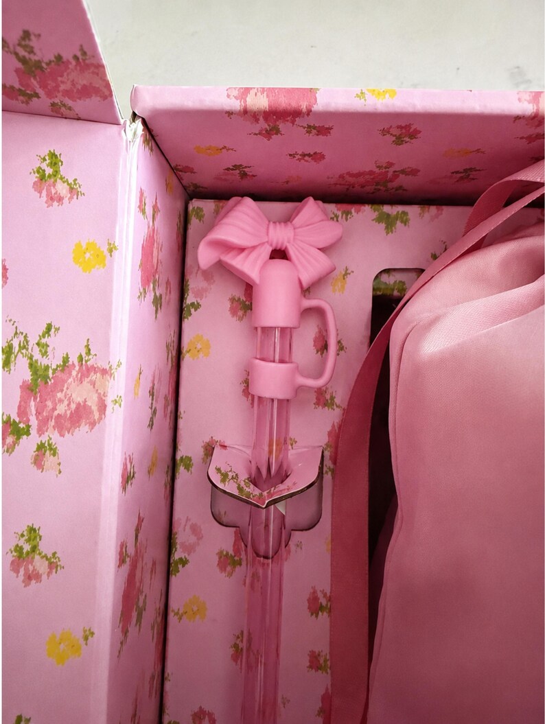 May include: A pink bottle stopper with a bow-shaped handle inside a floral-patterned box. The stopper has a long, clear tube and a small handle. The box and stopper are shades of pink, with a floral design.
