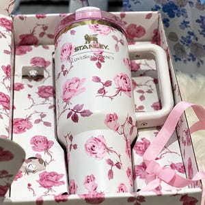 May include: White Stanley tumbler with a pink rose pattern, a pink lid, and a white handle. The tumbler is in a gift box with a matching floral design. The text "Stanley" and "LoveShackFancy" are visible on the tumbler.