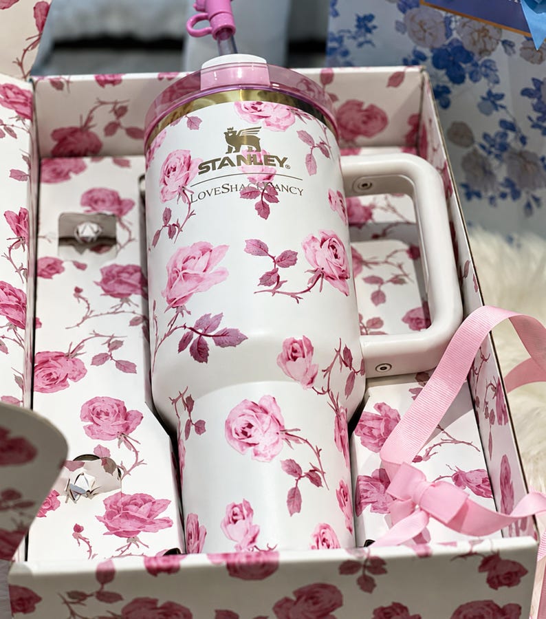 May include: White Stanley tumbler with a pink rose pattern, a pink lid, and a white handle. The tumbler is in a gift box with a matching floral design. The text "Stanley" and "LoveShackFancy" are visible on the tumbler.