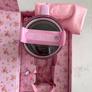 May include: A pink Stanley thermos with a matching lid and handle, nestled inside a floral-patterned box. A pink cloth with the Stanley logo is also included. The box and thermos are a soft pink color.
