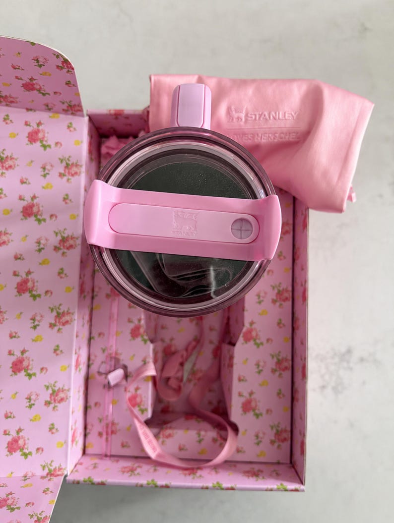 May include: A pink Stanley thermos with a matching lid and handle, nestled inside a floral-patterned box. A pink cloth with the Stanley logo is also included. The box and thermos are a soft pink color.