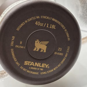 以下が含まれることがあります： 茶色のスタンレーウォーターボトルの底のクローズアップ。テキストには「Designed in Seattle WA」と「40oz / 1.18L」と書かれています。スタンレーのロゴは金色です。ボトルは食器洗い機対応で、BPAフリーです。