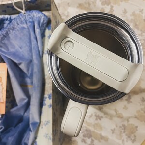 May include: A stainless steel tumbler with a clear lid and a white handle. The lid has a white handle with the Stanley logo. The tumbler is inside a box with a blue cloth.