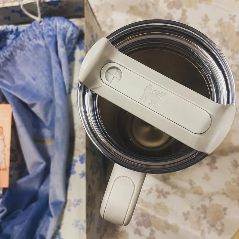 May include: A stainless steel tumbler with a clear lid and a white handle. The lid has a white handle with the Stanley logo. The tumbler is inside a box with a blue cloth.
