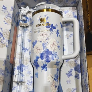 May include: A white Stanley tumbler with a floral design featuring white and blue flowers. The tumbler has a white handle and a gold-colored rim. The text "STANLEY LOVESHACK FAN" is printed on the tumbler. The tumbler is packaged in a box with a matching floral pattern.