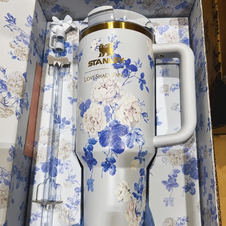May include: A white Stanley tumbler with a floral design featuring white and blue flowers. The tumbler has a white handle and a gold-colored rim. The text "STANLEY LOVESHACK FAN" is printed on the tumbler. The tumbler is packaged in a box with a matching floral pattern.