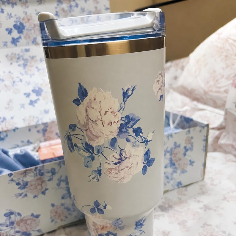 May include: A light gray tumbler with a floral design featuring white and pink roses and blue leaves. The tumbler has a gold rim and a clear plastic lid. A matching box is visible in the background.
