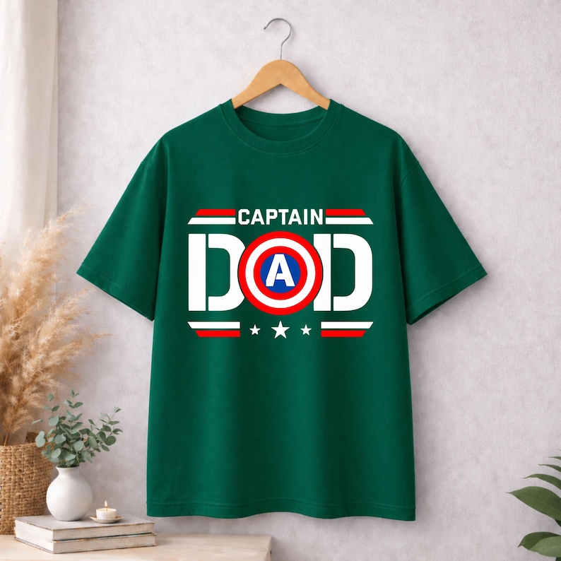 May include: A dark green short-sleeve t-shirt featuring a graphic print that reads CAPTAIN DAD in white letters. The letter O is replaced by a circular red, white, and blue shield design with a blue A in the centre, accented by red stripes and stars.