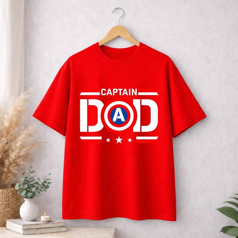May include: A bright red short-sleeved t-shirt featuring a white graphic print that reads CAPTAIN DAD. The letter O in DAD is replaced by a circular shield design with a blue centre and a white letter A, flanked by three small white stars.