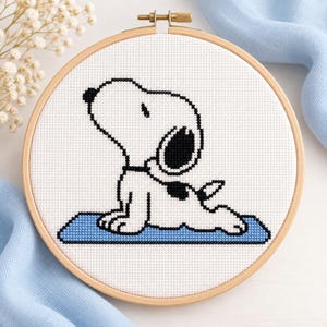 May include: A circular wooden embroidery hoop containing a white fabric cross-stitch pattern of a white dog with black ears and a black spot on its back, positioned in a yoga cobra pose on a rectangular blue mat, outlined in black thread.
