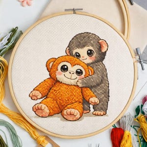 May include: Cross-stitch embroidery hoop featuring two cartoon monkeys. One monkey is orange and seated, while the other is gray and standing behind it. The monkeys have large eyes and are smiling. Embroidery floss in various colors surrounds the hoop.
