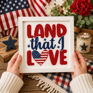 May include: A square framed cross-stitch embroidery featuring the red text LAND, the blue text that I, and the red text LOVE, with the letter O replaced by a heart patterned with the American flag, all on a white fabric background.