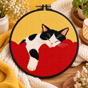 Può includere: Un motivo a punto croce raffigurante un gatto bianco e nero all'interno di un telaio da ricamo circolare nero. Il gatto è ritratto mentre dorme su una superficie rossa solida su sfondo giallo brillante, con dettagli cuciti di orecchie, baffi e zampe.