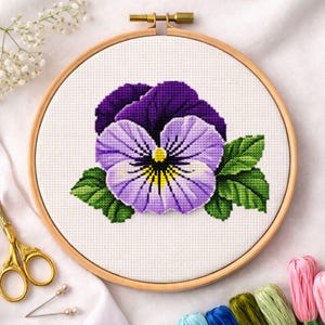 May include: A completed cross-stitch embroidery of a pansy flower in a wooden hoop. The pansy features purple, lavender, yellow, and green colors. The embroidery is on white fabric, and the hoop is secured with a brass clasp. Gold scissors and embroidery floss are visible.