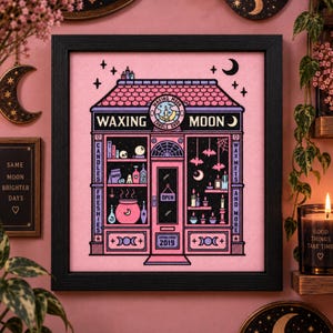 Peut inclure: Une broderie au point de croix encadrée représentant une devanture de magasin intitulée Waxing Moon Candle Shop. Le motif présente un bâtiment rose aux contours noirs, des étagères remplies de bougies, un chaudron et des chauves-souris. Texte inclus.