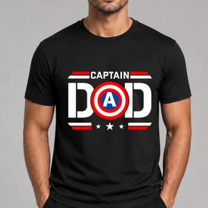 May include: A black short-sleeved t-shirt featuring a graphic print with the text CAPTAIN DAD in white block letters. The letter O is replaced by a circular red, white, and blue shield emblem with a blue A in the centre, flanked by red and white stripes.