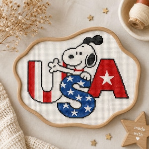 Może przedstawiać: Haft krzyżykowy w drewnianej ramce przedstawiający postać Snoopy'ego za literami USA. Litera U ma czerwono-białe pionowe paski, S jest niebieska w białe gwiazdki, a A jest czerwona z białą gwiazdą.