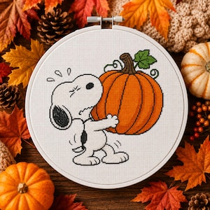 Può includere: Un progetto a punto croce su tessuto bianco inserito in un telaio da ricamo circolare in legno. Il disegno raffigura un cane dei cartoni animati in bianco e nero che fatica a sollevare una grande zucca arancione rotonda.