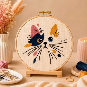 Puede incluir: Un bastidor de bordado circular de madera que contiene un patrón de punto de cruz de una cara de gato. El diseño presenta una paleta de colores azul marino, amarillo y rosa sobre tela crema, con bigotes negros bordados.