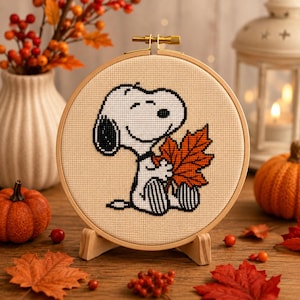 Peut inclure: Un tambour à broder circulaire en bois contenant un motif au point de croix du personnage de dessin animé Snoopy, blanc avec des oreilles et des marques noires, tenant une grande feuille d'érable orange et rouge sur un tissu beige clair.