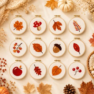 May include: Twelve circular embroidery hoops containing cross-stitch patterns of autumn motifs including various leaves, a mushroom, an acorn, a red apple, and berries. Each hoop features a white fabric background with detailed seasonal designs.