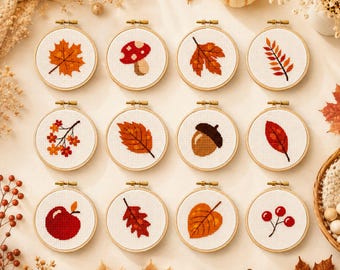 May include: Twelve circular embroidery hoops containing cross-stitch patterns of autumn motifs including various leaves, a mushroom, an acorn, a red apple, and berries. Each hoop features a white fabric background with detailed seasonal designs.