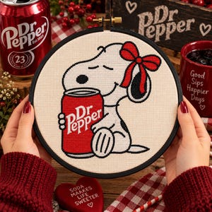 May include: A circular embroidery hoop containing a cross-stitch design of a white cartoon dog with a red bow on its head, holding a red soda can with the white text Dr Pepper printed on it. The design is set against a cream-colored fabric background.