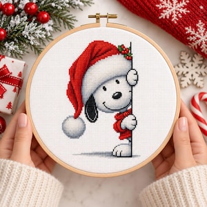 May include: A circular wooden embroidery hoop containing a cross-stitch pattern of a white dog with a black ear wearing a red Santa hat and a red sweater. The dog is peeking from behind a vertical line on a white fabric background.