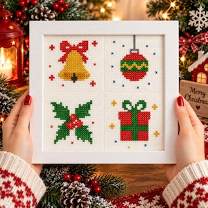 Op de afbeelding: Een vierkante witte lijst met een kruissteekborduurwerk met vier verschillende kerstmotieven: een gele bel met een rode strik, een rood-groen gestreepte kerstbal, een groene hulsttak met rode bessen en een rood cadeau met een groene strik.