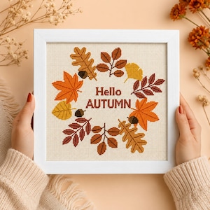 Può includere: Una cornice quadrata bianca contenente un ricamo a punto croce su tessuto color crema. Il design presenta una disposizione circolare di varie foglie autunnali nei toni dell'arancione, giallo e marrone, con ghiande e il testo Hello AUTUMN.