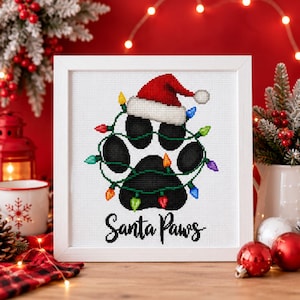 May include: A framed cross-stitch artwork featuring a black animal paw print wearing a red and white Santa hat, wrapped in a string of colorful Christmas lights, with the black text "Santa Paws" printed underneath the design.