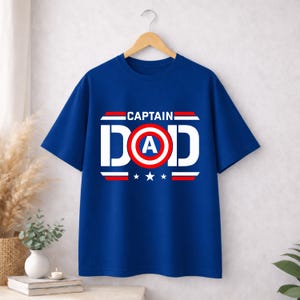 May include: A royal blue short-sleeve t-shirt featuring a graphic print that reads CAPTAIN DAD in white letters. The letter O is replaced by a red, white, and blue circular shield emblem with a white A in the centre, flanked by red and white stripes.