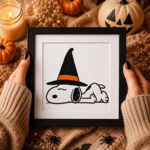 May include: A square black frame containing a cross-stitch embroidery of a white dog with a black ear, lying down and wearing a tall black witch hat with an orange band. The design is stitched on white fabric with a white mat inside the frame.