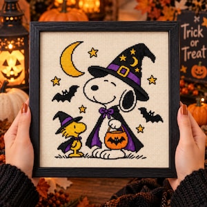May include: A framed cross-stitch embroidery featuring a white beagle wearing a purple and black witch hat and cape, holding an orange pumpkin bucket. A small yellow bird stands nearby in a matching hat and cape. The design includes a yellow crescent moon, stars, and black bats on a cream-colored fabric background.