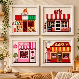May include: Four framed cross-stitch artworks depicting storefronts: a pizza shop with a bicycle, a cafe with a menu board, a pink candy shop with a lollipop sign, and a bakery with a chimney. Each square piece features detailed shop facades.