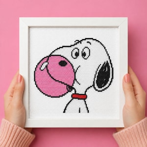 Può includere: Un ricamo a punto croce incorniciato raffigurante un cane bianco dei cartoni animati con un orecchio nero e un collare rosso, che soffia una grande bolla rotonda di colore rosa brillante. Il disegno è ricamato su tela Aida bianca in cornice.