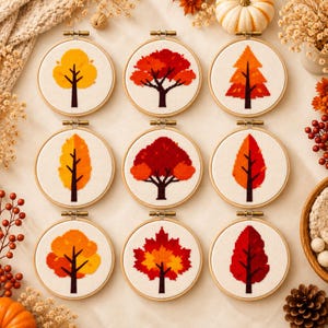 Könnte beinhalten: Neun runde Stickrahmen aus Holz mit Kreuzstichmotiven verschiedener Bäume. Jeder Rahmen zeigt einen Baum mit herbstlich gefärbtem Laub in Gelb-, Orange- und Rottönen auf einem weißen Stoffhintergrund mit einem sichtbaren Rastermuster.