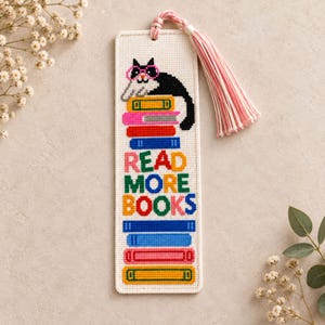 Puede incluir: Un marcapáginas rectangular de punto de cruz sobre tela color crema con un gato blanco y negro con gafas rosas posado sobre una pila de libros coloridos. El texto READ MORE BOOKS está bordado en letras multicolores y tiene una borla rosa.