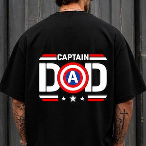 May include: A black short-sleeved t-shirt featuring a graphic on the back with the text CAPTAIN DAD in white letters. The letter O is replaced by a circular shield design in red, white, and blue with a white letter A in the centre.