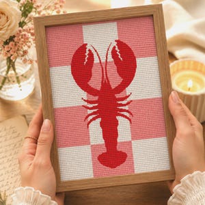 May include: A framed cross-stitch embroidery featuring a vibrant red lobster centered against a background of alternating white and light pink checkered squares. The artwork is encased in a simple, light-toned wooden frame with a clean finish.