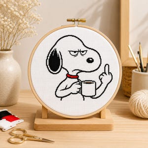 May include: A circular wooden embroidery hoop containing a cross-stitch design of a white cartoon dog with a black ear and red collar. The dog has a grumpy expression, holds a white coffee mug, and is gesturing with its middle finger raised.